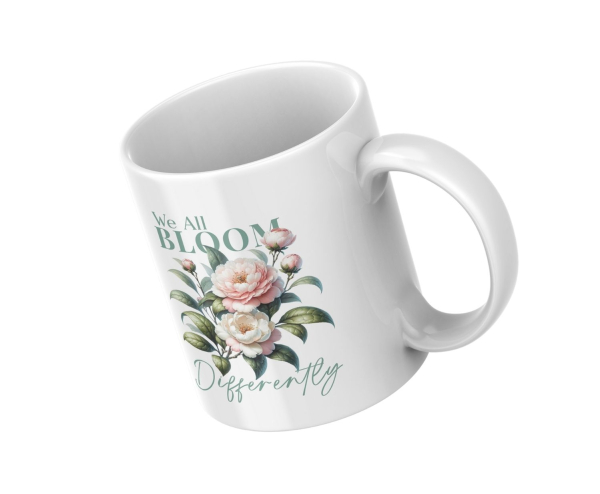Weiße Keramiktasse mit rosa Blüten und dem Spruch „We All Bloom Differently“, schräg von oben fotografiert auf weißem Hintergrund.
